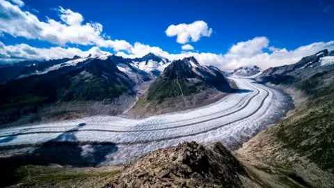 Getty Images View dari Aletsch Glacier. Es menyapu bulat dari kanan ke kiri, dengan pegunungan di kedua sisi. Di atas adalah langit biru dengan beberapa awan.