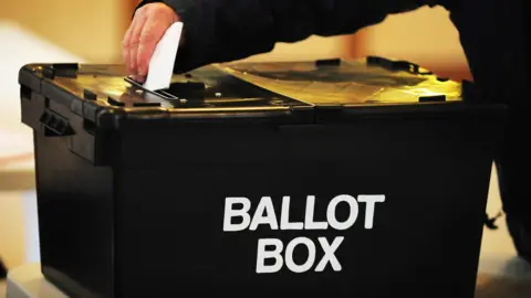 Hand placing a while ballot in to a black ballot box with the words 'ballot box' written on it in white. 