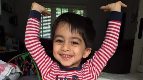 A three-year-old boy wearing red and white striped pyjamas grins at the camera. His arms are above his head and a typical family living room is visible behind him. 