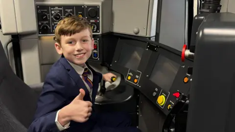 A young boy with brown hair and a blue suit with one thumb in the air and the other hand on a lever in the cab of a train with various buttons, monitors and levers around him