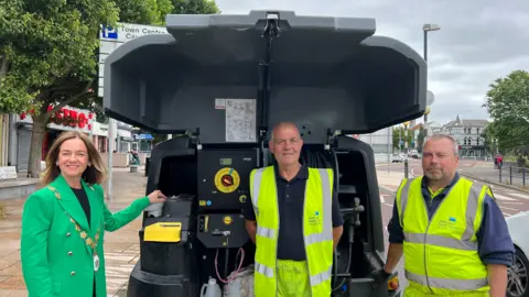 Councillor Gillian McCollum stands in a green jacket beside Paul Agnew and George Irwin who both wear high vis jackets. They stand in front of a street washing machine.