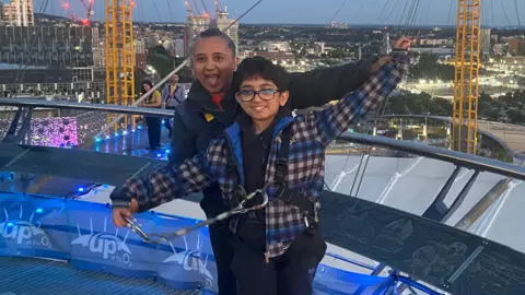 Rajeshree Shah stands with her child Aadi on top of the 02 arena, smiling and waving.