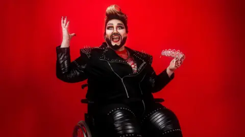 Drag artist Emma Round sitting in a wheelchair while wearing a black leather biker jacket and trousers. They are holding a tiara and sitting in front of a red backdrop. 