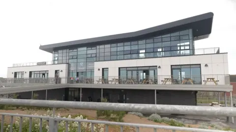 A large, modern building with white cladding, large glass windows and a sweeping black roof. The Seaview Colonnade has a terrace around it, with picnic benches behind metal railings. The building is raised off the ground, with a large raised ramp up to the entrance. There is sand and beach plants in the foreground.