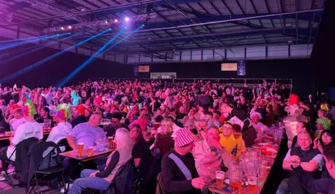 People are sitting at tables, which have beer glasses on them. The bottom half of the image is packed with people, some in hats and fancy dress, with the ceiling above them. 