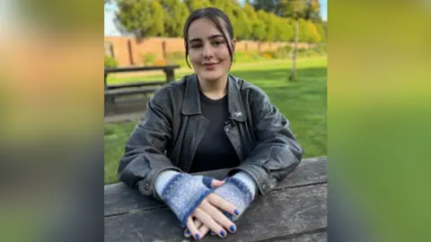 Manon Chiswell está mirando la cámara. Ella está sentada en una mesa de picnic con la mano sobre la mesa misma. Ella tiene barniz de uñas azules y guantes azules sin dedo. Lleva una chaqueta de cuero negra debajo de una camiseta negra.