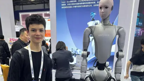 Nathan Lepora Jared at a robotics convention, wearing a black top and convention white lanyard. He is standing next to a white robot. In the background there are other attendees and other robotics equipment.