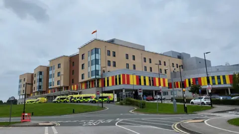 BBC A hospital with ambulances parked outside it. The hospital building is several storeys high, and has red and yellow cladding on the lower part.
