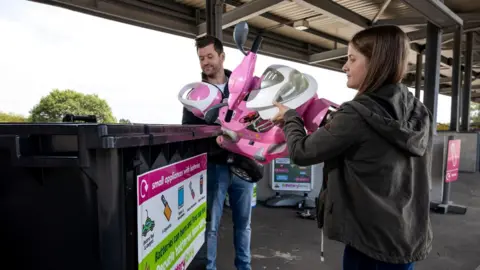 A man and woman, both with brown hair, lift a child's pink electric moped into a large rubbish bin