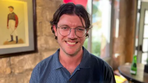 Harry Harvey is pictured wearing a dark blue shirt and round glasses. He is smiling widely directly at the camera. He has short dark hair parted in the middle and a light-coloured moustache. He is standing in front of a stone wall in a cafe.
