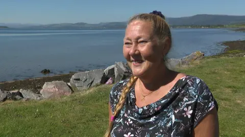 Maria, standing outside on the shores of the Cromarty Firth, has her blonde hair tied in a long braid. She is wearing a dark top with a colourful floral pattern. Maria is smiling, and the sun is shining.