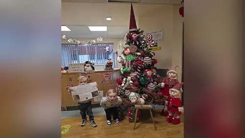 Family photograph Left: Lucas, three, holds up a poster covering his lips about Parent Support Boxes with Leo to his right bending down with his hands in his pockets and mouth wide open. They are standing next to a Christmas tree with decorative elves on the right in the reception of a children's ward in a hospital.
