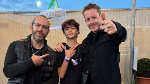 Family handout A young boy in a black T-shirt posting for a photo with two men, both wearing black jackets. They're standing in front of a temporary festival fence and the sky is blue.