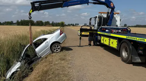 A large silver car is being winched from a ditch at the side of a road. A man can be seen near the truck, guiding the mechanism