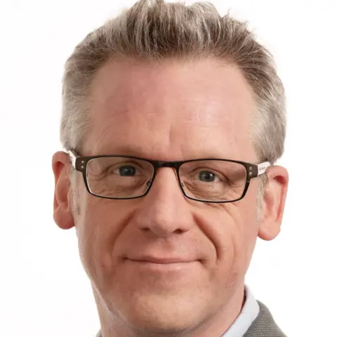 Mark Miller A man is looking at a camera with a white background behind him. He is in a shirt and blazer and is wearing glasses.