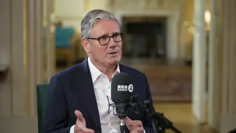 Sir Keir Starmer sits in an elegantly decorated room in Downing Street, wearing a dark suit and white shirt, gesturing with his hands as he talks. A BBC Radio 4 microphone is placed in front of him. The background includes a fireplace and ornate furnishings under soft lighting.
