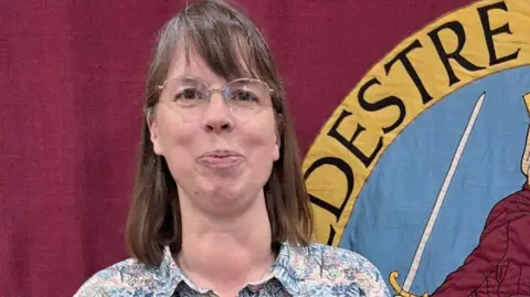 A woman with shoulder length brown hair and wire-framed glasses stands in front of a large town flag in maroon with part of a yellow, black and blue crest visible.