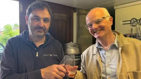 Two men pose for a photo, smiling, while clinking two glasses with a small amount of ale at the bottom. Kevin, on the left, is wearing a dark blue zipped-up hoodie and has a black and grey beard and black short straight hair. David, on the right, has white thinning hair, glasses and is wearing a white and blue patterned shirt and beige light jacket over the top. Beer kegs can be seen behind them.