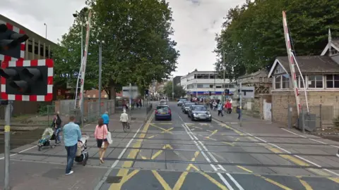 Google Eight pedestrians crossing a level crossing near a train station. There are yellow road markings and cars on both sides of the road in the background.