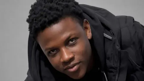A head shot of Panashe Muchenje who is a black man with black hair. He is smiling and wearing a black hoodie.