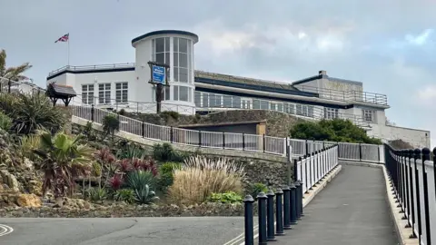 An exterior view of the art deco Winter Gardens building. It is a white building constructed of various shapes, with dirt on some walls. It sits on the cliff edge with a long path leading up to it.