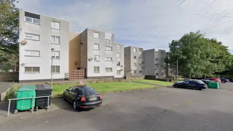 Google A row of white 1970s flats with some care parked in front of them.