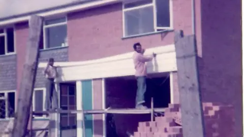 Dr Yana Johnson An old image of two men fixing up the front of a house. There are pieces of wood and piles of bricks in the foreground.