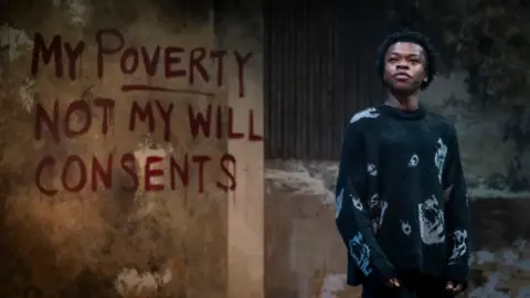 Belgrade Theatre Kyle Ndukuba dressed in a black jumper in stood in a dilapidated warehouse with the words 'my poverty not my will consents' spray painted on the wall