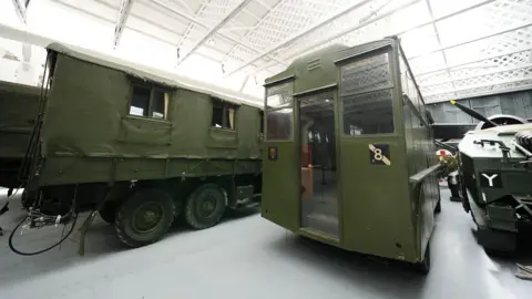 PA Media Three green army like caravans are stood in a hangar at a museum. One of them, just off centre to the right, has a large glass door and two glass windows either side. All of them have several wheels.