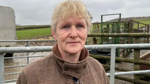 Carol Field, who has blonde hair and blue eyes, is looking at the camera. She is wearing a thick brown coat and is on a farm, with gates, machinery and a field behind her.