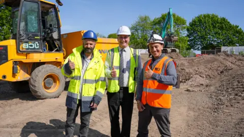 Three men in hi-vis jackets and hard hats stand together on a building site, looking at the camera and smiling with their thumbs up. On the building site behind them is a yellow digger and mounds of dirt.