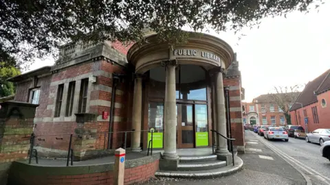 BBC An old building with a sign saying Public Library at the top. It is a listed building with red bricks and an automatic door in front. It is on the edge of a pavement with traffic queueing on the road