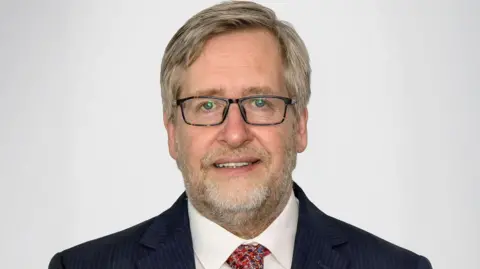 Scottish Care Man wearing a dark pinstripe suit with a white dress shirt and a patterned red tie, posed against a plain light gray background.