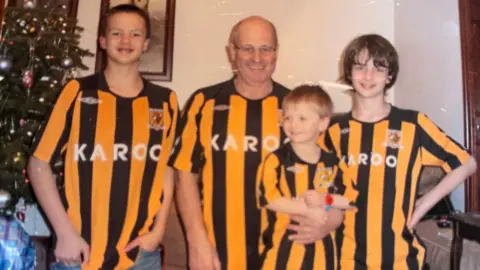 Jasmine Lowe/BBC News An old photograph of a younger Geoff and his grandsons as children next to a Christmas tree. They are all wearing identical Hull City shirts. Geoff has his arm around Jacob. 