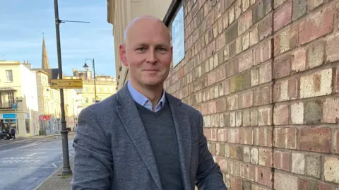 Max Wilkinson smiles as he stands in front of a brick wall in Cheltenham town centre. There are Regency style shop fronts behind him and a church spire, which are glowing in the winter sun. He is wearing a dark grey blazer over a dark grey jumper and a blue shirt collar.
