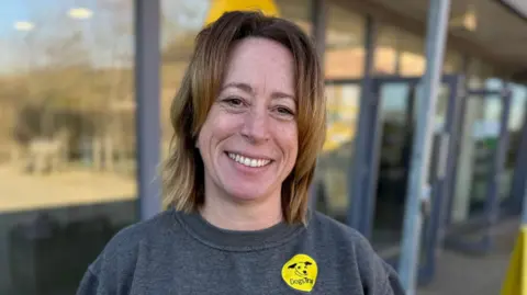 Lisa Cooper, who has shoulder-length brown hair, smiling at the camera. She is standing in front of windows and is wearing a dark grey sweater that has a yellow Dogs Trust sticker on it.