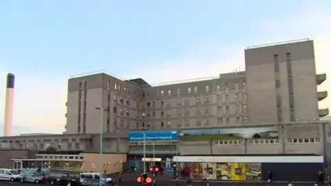 The grey building of Derriford Hospital in Plymouth, also seen are people walking around and a pedestrian crossing in front of the hospital, as well as a number of vehicles parked up outside