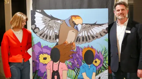 A woman and man stand either side of a collage painting showing two children sitting in a field with a falcon above them and purple flowers behind. Their faces are cut out so people can stand in put their own faces in the gap. The woman has a red cardigan and blue jeans, and the man is wearing a suit jacket, jeans and a checked white shirt.