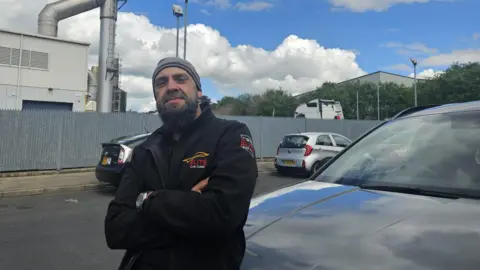 Aisha Iqbal/BBC A man stands in front of what looks like a factory plant on a road with cars parked around him. He leans against a car, arms folded.