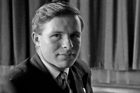 King Edward's School A black and white photo of a boy in a school blazer and striped tie with short, brushed hair. He is sitting in front of a drawn dark curtain and looking at the camera.