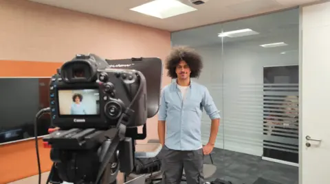 Phil Hocking A man in the background being filmed by a camera on a tripod in the foreground. He is visible on the screen in the camera and the wall on the left of the photo is yellow.