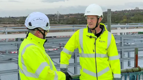 Two men are standing next to each other on a building site wearing white hard hats and high vis jackets which say 'agratas'