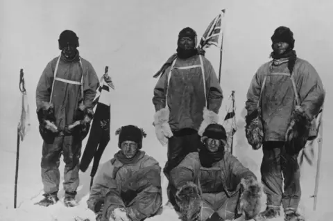 Getty Images Black and white image of Captain Scott, Edward Wilson, Henry Bowers, Lawrence Oates and Edgar Evans at the South Pole. Three men are standing, and two are sitting on the snow in a posed photograph. They are wearing warm clothing but look cold and exhausted. Several flags have been planted.