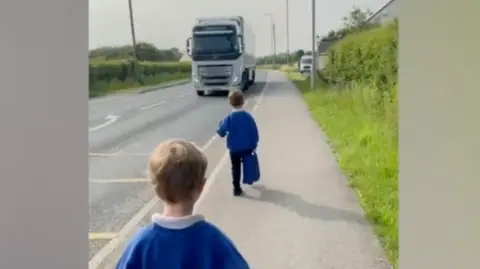 Two children in blue school jumpers walking down the road towards the primary school about a metre away from a lorry driving down a main road