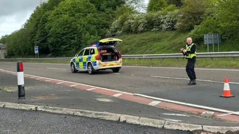 A police officer looks across the A1 in Northumberland. A police car with an open boot is parked in the middle of the closed road.