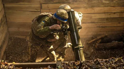 Getty Images Two Ukrainian soldiers lean over mud in a wooden bunker preparing to fire at Russian positions in the Sumy region on September 30, 2024.