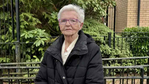 Anna Stuart, an older lady with short white hair and purple glasses. She looks seriously into the camera. She wears a black quilted anorak and a cream-coloured blouse.