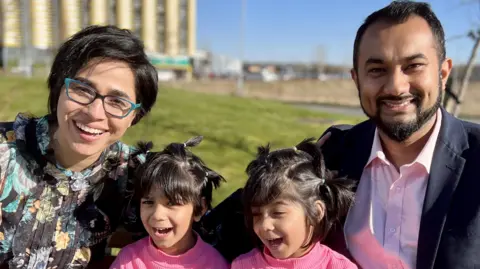 Sultana smiles at the camera next to her husband Nayeem Ahmad. Their twin girls sit in between them wearing matching pink tops.