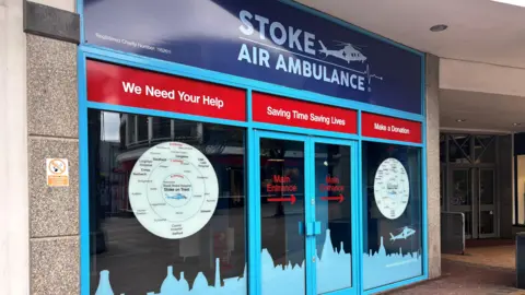 The front of a shop which has large windows with blue vinyl attached with logos and information about the Stoke Air Ambulance charity.