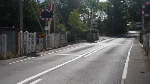 Stuart Woodward/BBC A wide shot of a level crossing which is currently open for traffic to pass through. Trees are in the background.